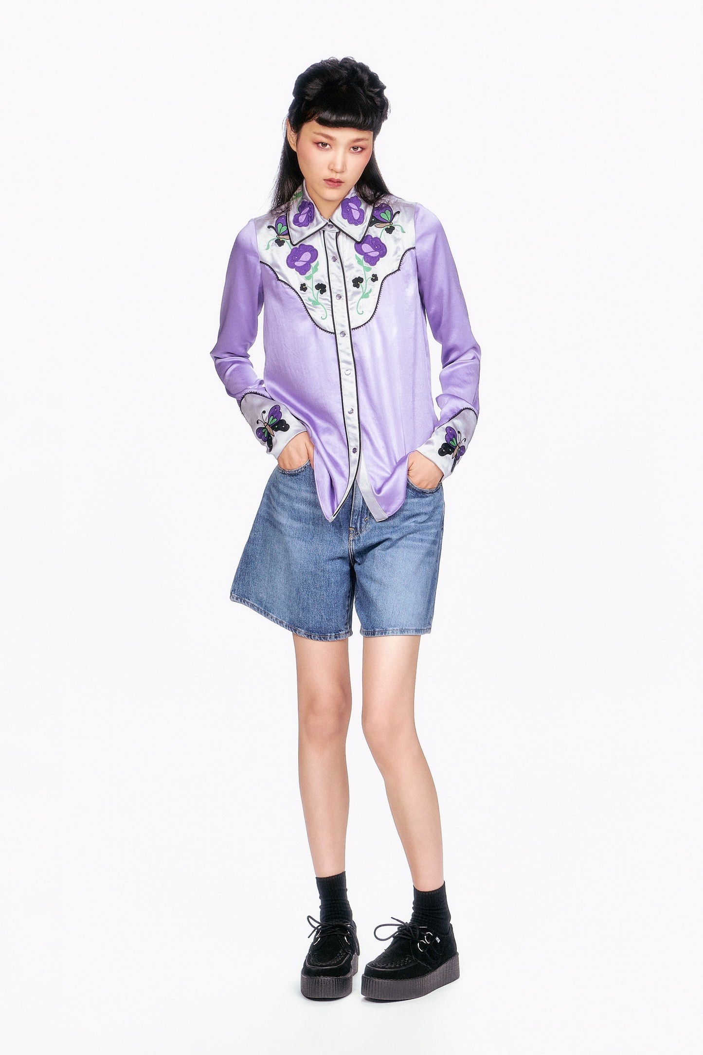 Person wearing a light purple shirt with floral embroidery waist long and denim shorts