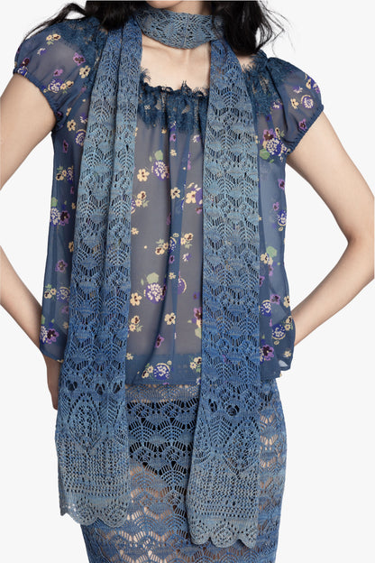 Woman wearing a blue floral short sleeve top, blue gradient crochet scarf and matching skirt on a white background.