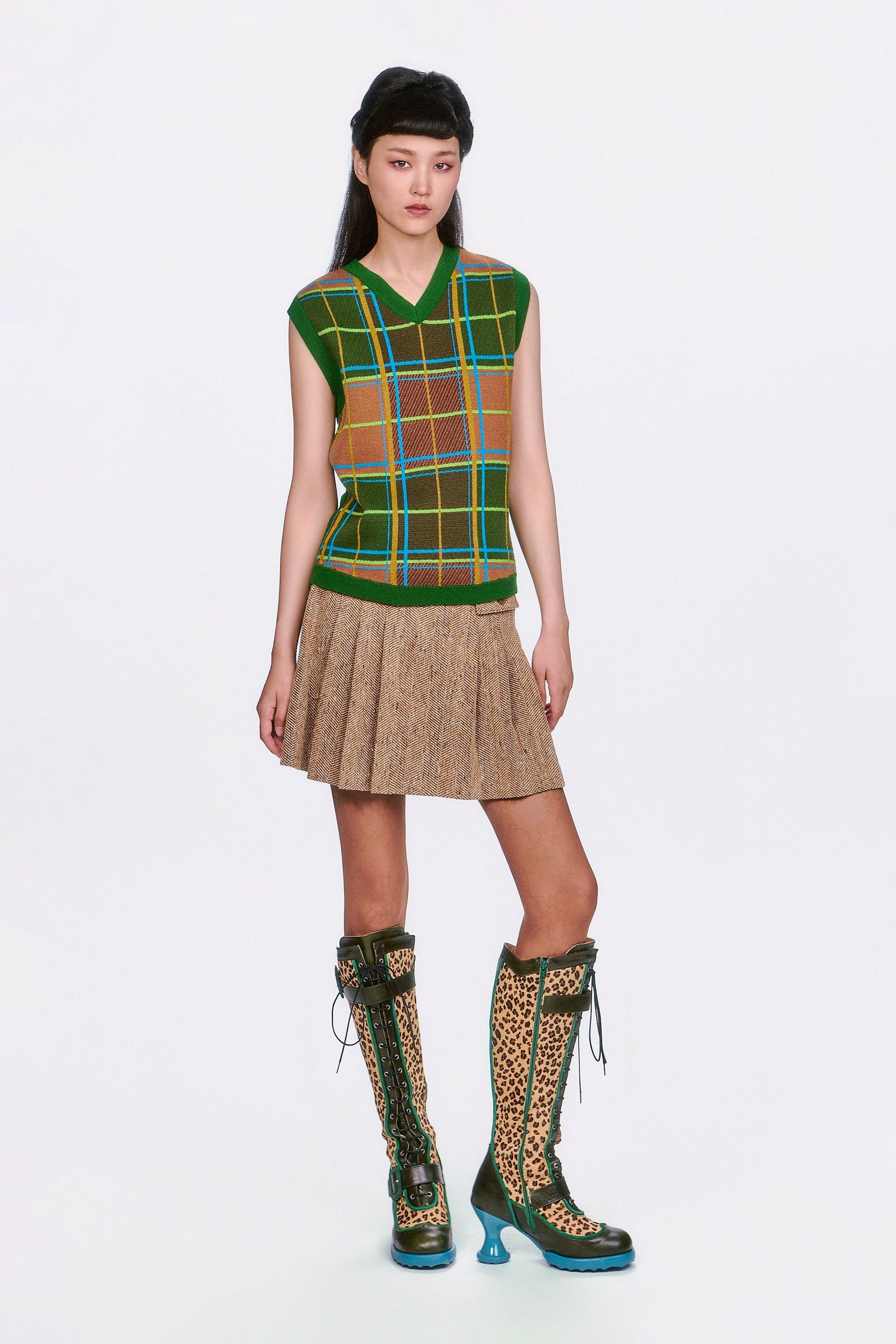 Model wearing a plaid sleeveless top with Herringbone Pleated Skirt Caramel with green knee-high boots. 
