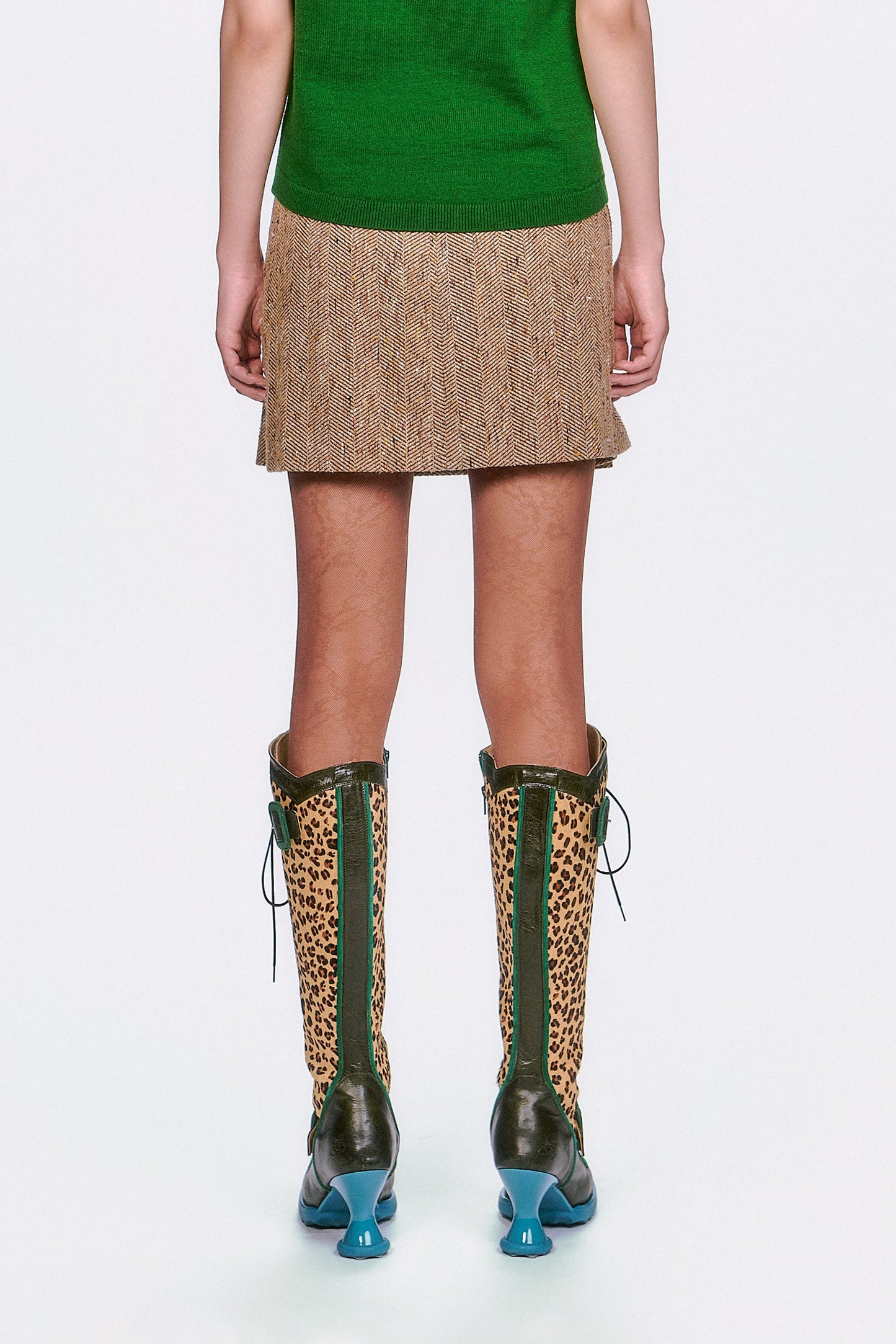 A woman wearing The Herringbone Pleated Skirt Caramel mid-tight high, green top & leopard print boots