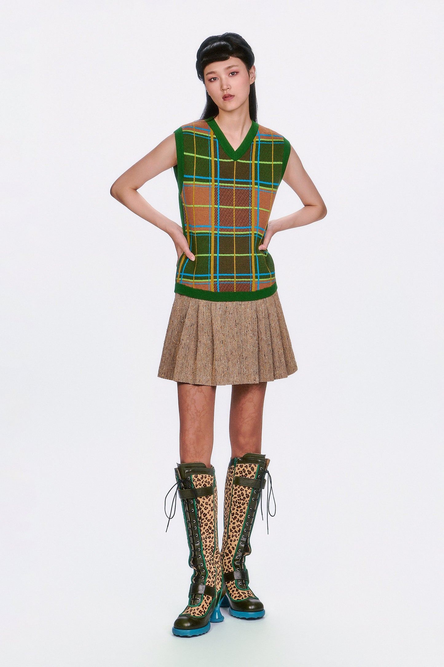 Model wearing a plaid sleeveless top with Herringbone Pleated Skirt Caramel with green knee-high boots. 