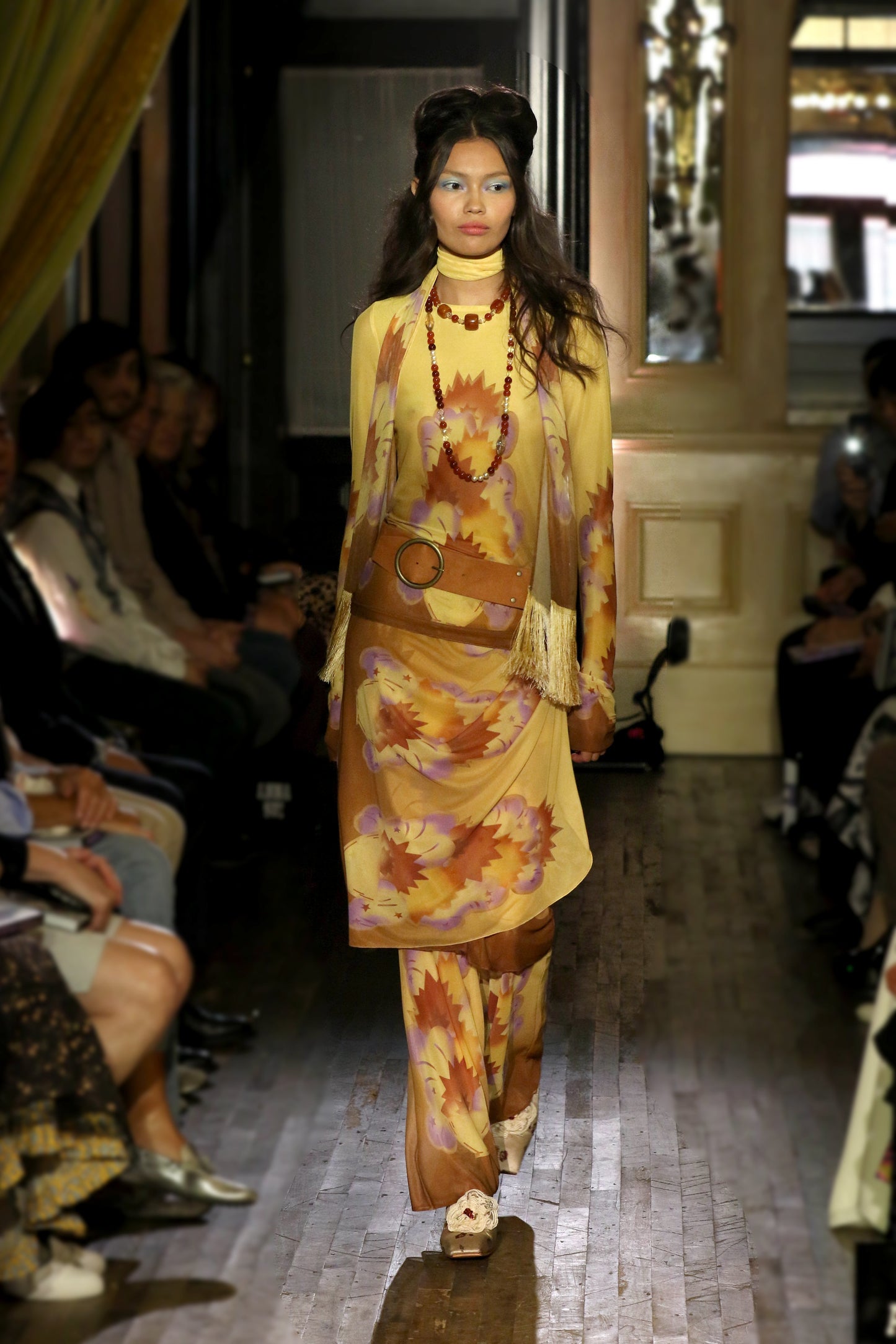 Model walking on a runway wearing a yellow geometric mesh outfit with a tan suede belt with audience watching.