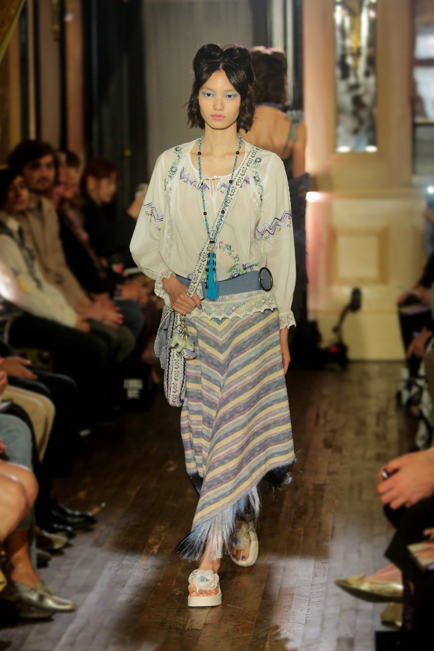 Model walking down runway wearing a white cotton embroidered top, blue belt, blue knit stripe skirt with fringe, long blue tassel necklace and bag, while audience watches.