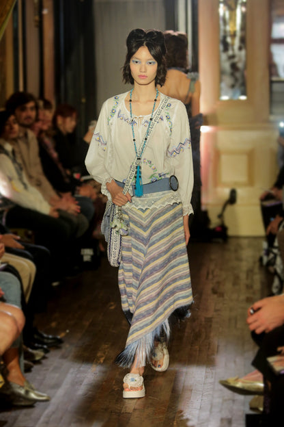 Model walking down runway wearing a white cotton embroidered top, blue belt, blue knit stripe skirt with fringe, long blue tassel necklace and bag, while audience watches.