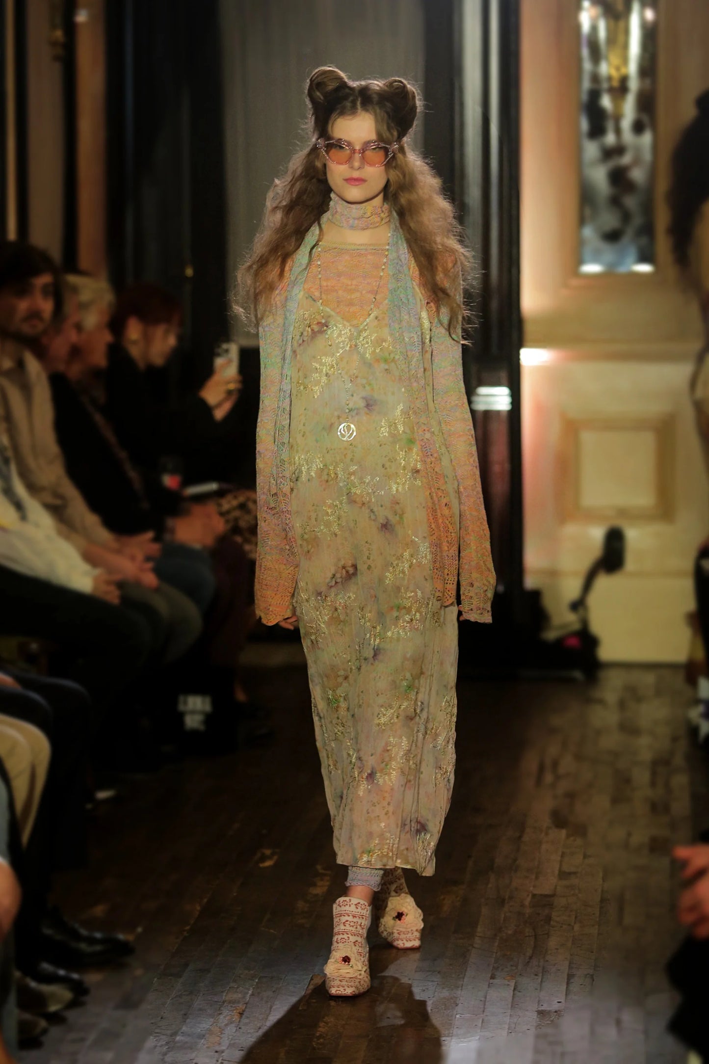 Model walking on a runway wearing a floral dress, crochet gradient long sleeve top and matching scarf, with an audience watching.