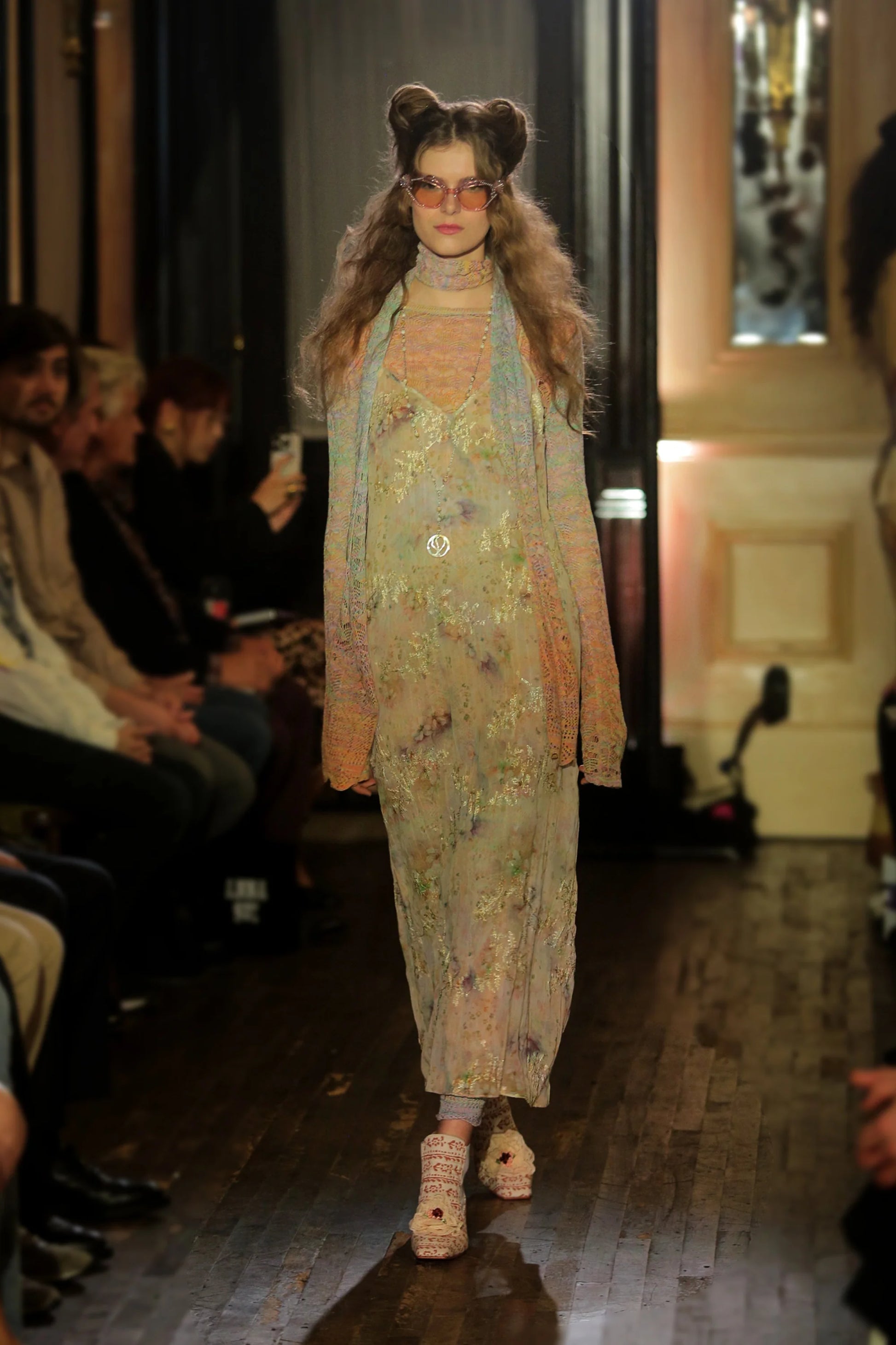 Model walking on a runway wearing a floral dress, crochet gradient long sleeve top and matching scarf, with an audience watching.
