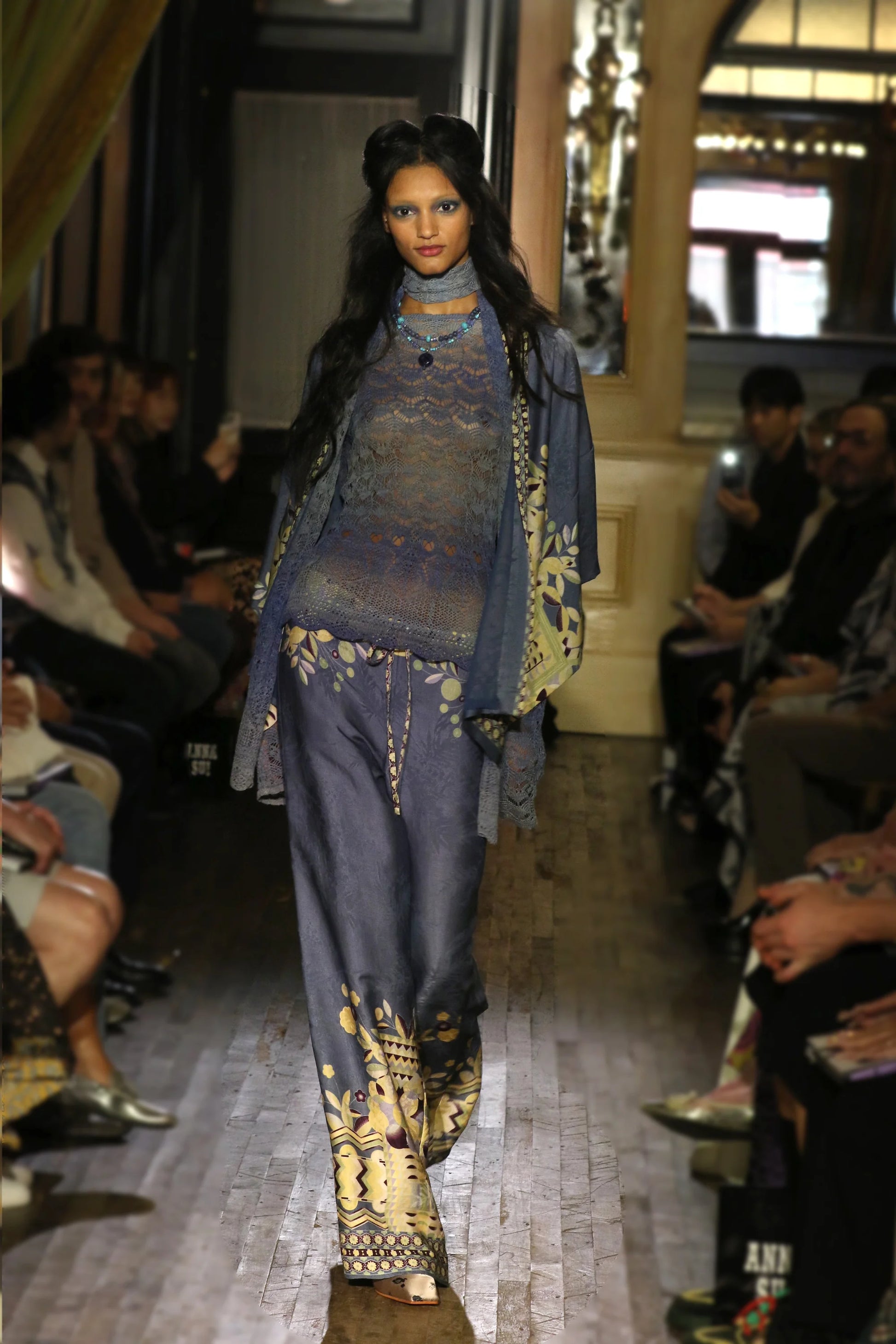 Model walking on a runway wearing a blue and gold outfit with a patterned scarf.