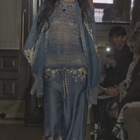 Video of a model walking on a runway wearing a blue and gold outfit with a patterned scarf with the audience watching.