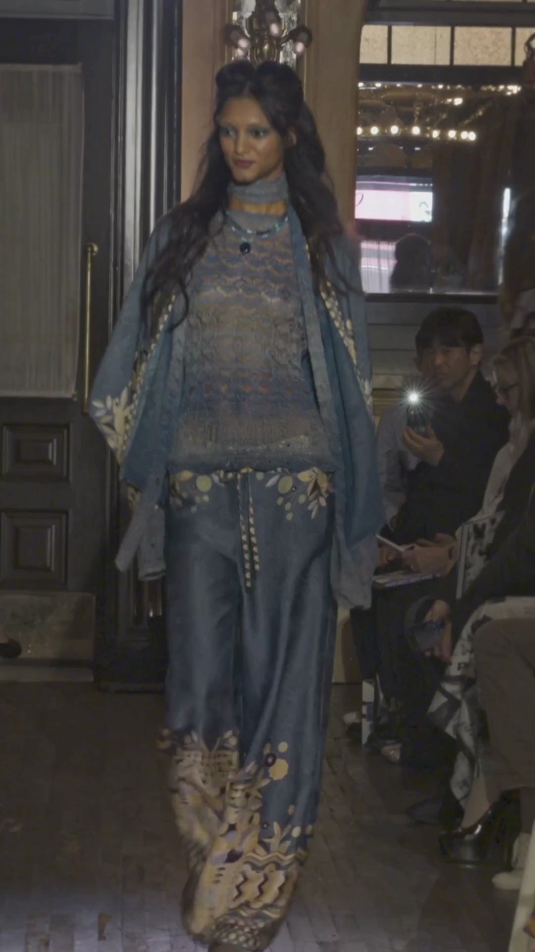 Video of a model walking on a runway wearing a blue and gold outfit with a patterned scarf with the audience watching.