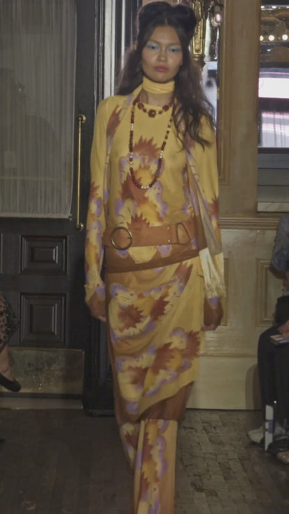 Model walking on a runway wearing a yellow geometric mesh outfit with a tan suede belt with audience watching.