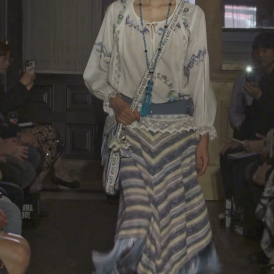 Video of model walking down runway wearing a white cotton embroidered top, blue belt, blue knit stripe skirt with fringe, long blue tassel necklace and bag, while audience watches.