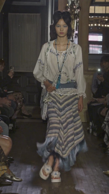 Video of model walking down runway wearing a white cotton embroidered top, blue belt, blue knit stripe skirt with fringe, long blue tassel necklace and bag, while audience watches.