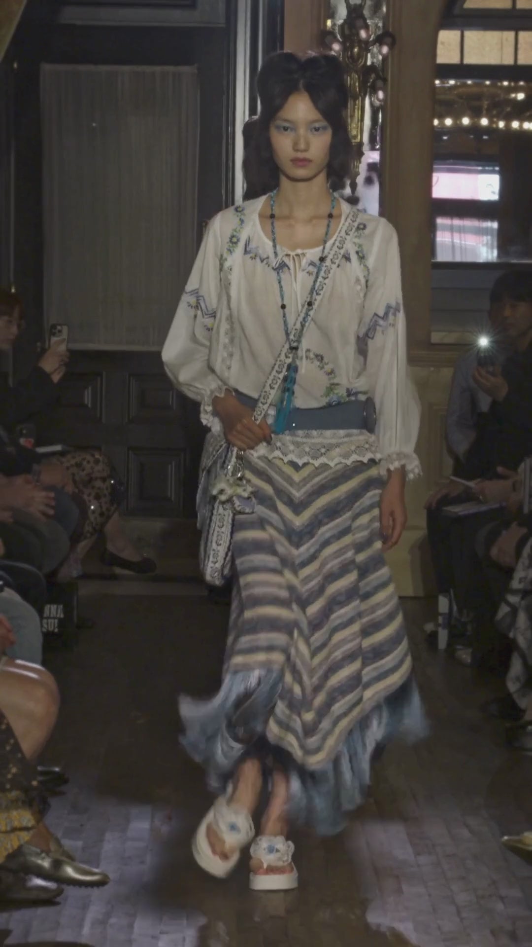 Video of model walking down runway wearing a white cotton embroidered top, blue belt, blue knit stripe skirt with fringe, long blue tassel necklace and bag, while audience watches.