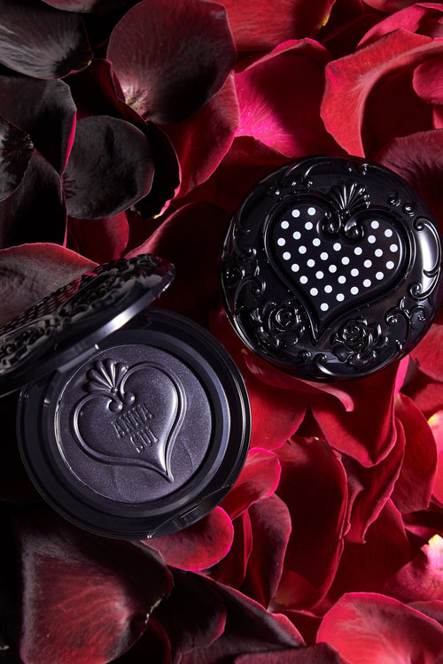 2-Cream blush in round black container, 1 open 1 closed, on a bed of red roses, top lid shows an engraved hart with white dots in it